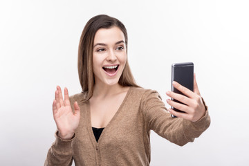 Fototapeta premium Perfect shot. Charming young woman posing for the camera, waving at it while taking selfies, standing isolated on a white background