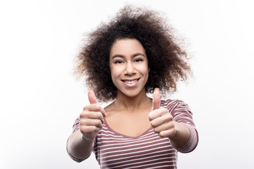Well done. Adorable young woman in a striped pullover showing thumbs up and smiling while posing for the camera, standing isolated on a white background