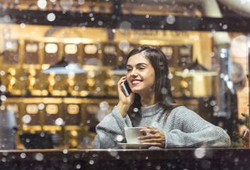 Young pretty brunette girl wears grey sweater have pleasant conversation by smartphone sitting near cafe window in snowy Christmas time