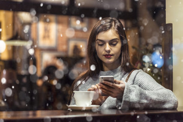 Young pretty brunette girl wears grey sweater holding smartphone sitting near cafe window in snowy Christmas time