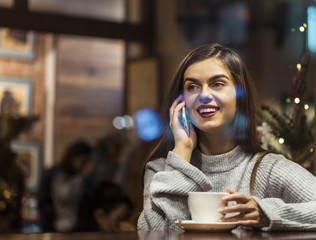 Young pretty brunette girl wears grey sweater have pleasant conversation by smartphone sitting near cafe window
