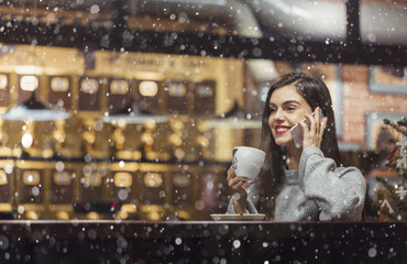 Young pretty brunette girl wears grey sweater holding smartphone sitting near cafe window in snowy Christmas time