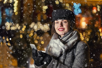 Stylish brunette smiling girl using smartphone before garland wall in city snowy evening Christmas time