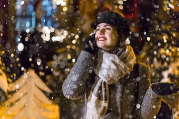Stylish brunette smiling girl having pleasant talk by smartphone before garland wall in city snowy evening Christmas time