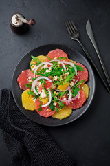  Orange, grapefruit, ricotta cheese, pine nuts, red onions and arugula with olive oil in ceramic plate on black concrete or stone old background. Selective focus. Top view. Copy space.