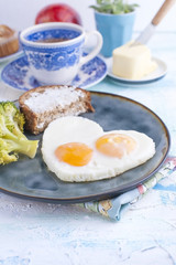 Fried eggs in the form of heart. On a dark plate with broccoli and toast. Blue cup with tea, butter for breakfast and cupcakes. On a light colored background. Free space for text or advertising.