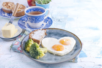 Fried eggs in the form of heart. On a dark plate with broccoli and toast. Blue cup with tea, butter for breakfast and cupcakes. On a light colored background. Free space for text or advertising.