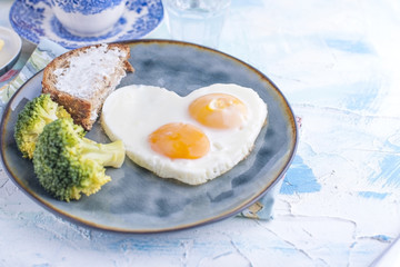 Fried eggs in the form of heart. On a dark plate with broccoli and toast. Blue cup with tea, butter for breakfast and cupcakes. On a light colored background. Free space for text or advertising.