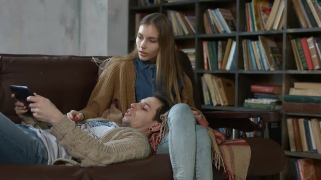 Handsome hipster lying on the couch with his head on beautiful girlfriend's lap and browsing the internet together using digital tablet at home. Young couple with tablet pc spending leisure together.