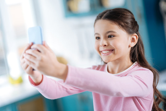 Joy. Pretty alert dark-eyed dark-haired schoolgirl smiling and taking selfies while holding her phone in front of her
