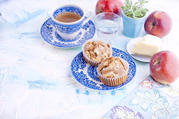 Homemade muffins with almonds on a blue plate for breakfast and tea in a cup. A glass of water and a red fresh apple and butter for sandwiches. Free place for text or postcard.