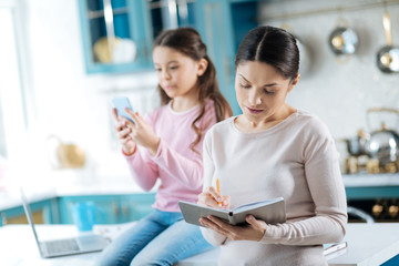 Nice atmosphere. Beautiful concentrated dark-haired woman holding and writing in the notebook while her daughter using her phone in the background