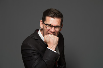 Emotional businessman in formal suit and glasses shows fist in camera threatening on gray background