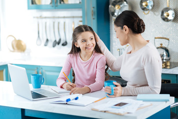 Obraz premium Caressing. Pretty alert dark-haired schoolgirl writing in her notebook and smiling at her mother standing near the girl with a cup of tea and caressing her