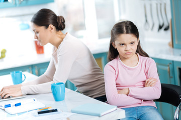 Fototapeta premium Boredom. Sweet dissatisfied dark-eyed girl having her arms crossed and sitting near her mom working on her laptop