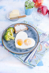 Fried eggs in the form of heart. On a dark plate with broccoli and toast. Blue cup with tea, apples, butter for breakfast. On a light colored background. Free space for text or advertising.