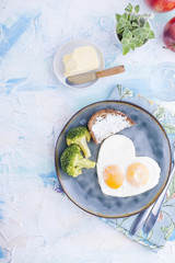Fried eggs in the form of heart. On a dark plate with broccoli and toast. Blue cup with tea, apples, butter for breakfast. On a light colored background. Free space for text or advertising.