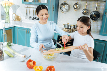 Healthy salad. Pretty inspired dark-haired girl smiling and putting ingredients into a bowl and making a salad with her mother