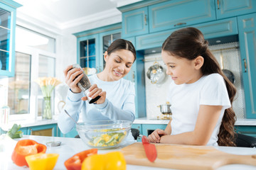 Healthy food. Beautiful inspired dark-haired mother peppering her salad and her daughter watching her and smiling