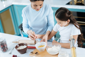 Great bakers. Attractive content dark-haired girl and her mother smiling and beating up some eggs while making dinner
