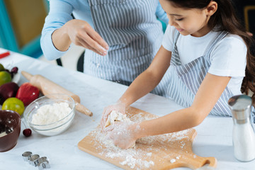 Fototapeta premium Funny baking. Lovely delighted dark-haired daughter making some dough and smiling and her mom helping her