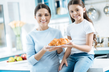 Happiness. Good-looking exuberant dark-haired little girl and her mother smiling and holding a plate with some croissants while the girl sitting on the table