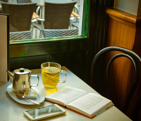 Glass Cup with Freshly Brewed Hot Green Tea Pot Honey Smartphone Open Book on Table by Window in Cafe. Wooden Chair Tranquil Romantic Cozy Atmosphere. European Style. Lifestyle Photo