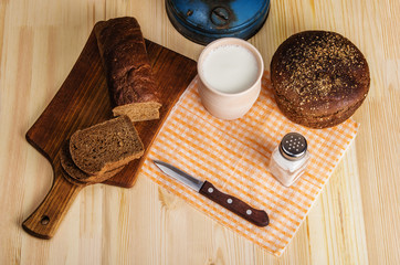 Fototapeta na wymiar Cutting board, clay pot with milk, rye bread and a knife on a wooden table. Still-life in a rustic style