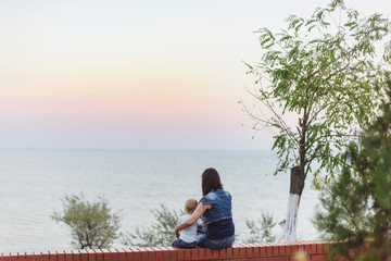 Young woman sitting on bench, looking at sea scarlet sunset, hugging little cute child boy on nature, green trees. Mother, little kid son. Parenthood, family day 15 of may, parents, children concept.