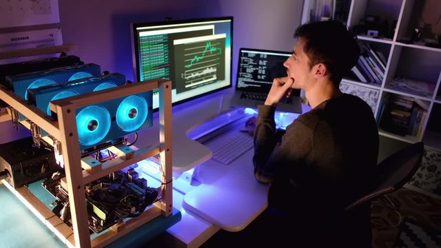 Cryptocurrency mining rig running with the young man controlling mining process and checking charts on the background in the evening at home.