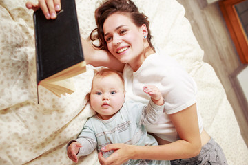 Young woman holding a book while laying with baby on a bed