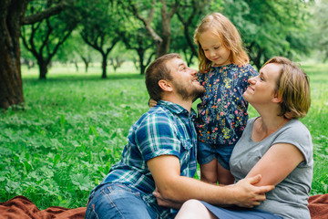 Fototapeta premium Young happy family of three lying on blanket in the park having fun. Happy parenting concept. Little girl with mother and father outdoors