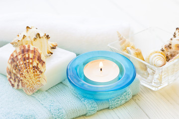 Aromatherapy Spa Concept with a fragrant candle in a blue candle holder, a bar of soap, terry towels, sea shells in glass clear bowls on white wooden background