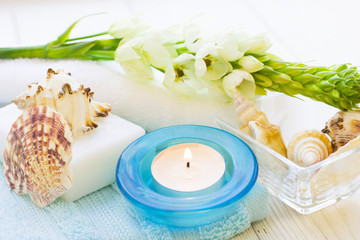 Aromatherapy Spa Concept with a fragrant candle in a blue candle holder, a bar of soap, terry towels, sea shells and white flower on white wooden background