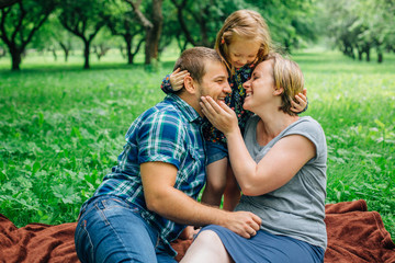 Fototapeta premium Young happy family of three lying on blanket in the park having fun. Happy parenting concept. Little girl with mother and father outdoors
