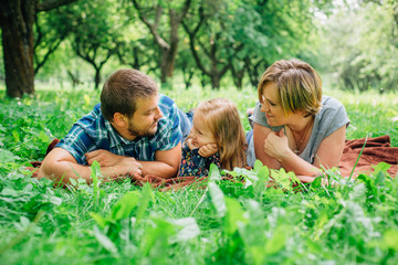 Young happy family of three lying on blanket in the park having fun. Happy parenting concept. Little girl with mother and father outdoors