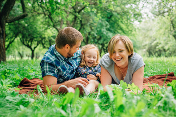Fototapeta premium Young happy family of three lying on blanket in the park having fun. Happy parenting concept. Little girl with mother and father outdoors