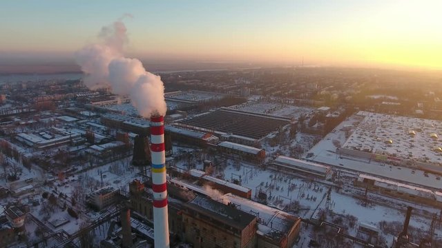 A bird`s eye view of a high cooling chimney with a slow flux of smoke at a splendid sunset in winter. It stands among snowy houses. 