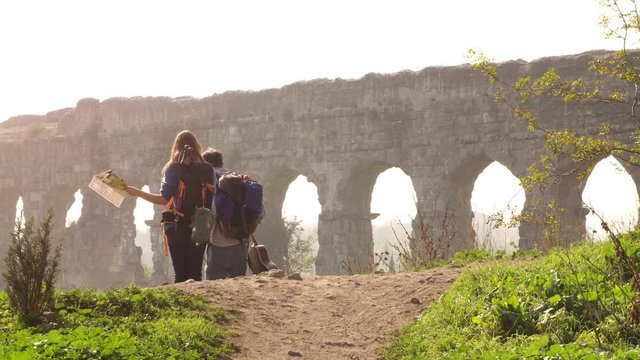 Young lovely couple backpackers tourists walking holding hands toward roman aqueduct arches in parco degli acquedotti park ruins in rome on romantic misty sunrise with guitar and sleeping bag slow