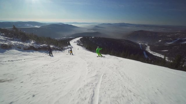 Sportsman on board in bright green clothes is riding down upon a slope of the hill at sunny winter day