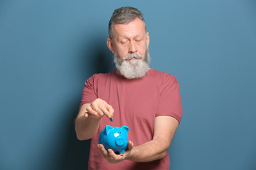 Senior man putting coin into piggy bank on color background