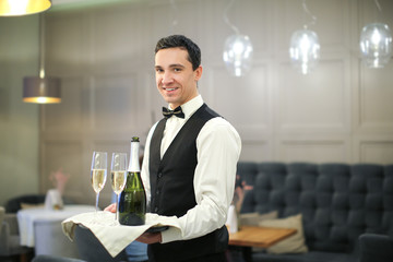 Young waiter holding tray with glasses and bottle of champagne indoors