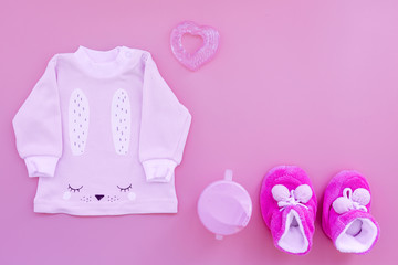 Cute pink baby clothes for girl. Shirt, booties, toy, bottle on pink background top view copy space