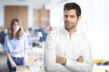 Fototapeta na wymiar Young professional man. Confident young financial assistant businessman wearing shirt and looking at camera while standing at the office. Unrecognizable businesswoman standing at the background. 