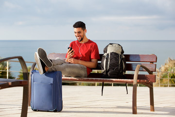 Fototapeta premium happy young man sitting on bench with luggage and listening to music with mobile phone and earphones