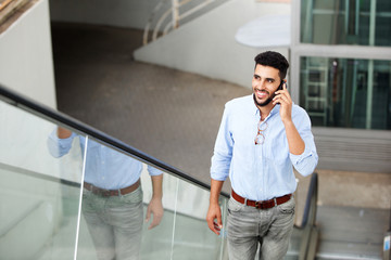 smiling young arabic man standing on escalator talking on mobile phone