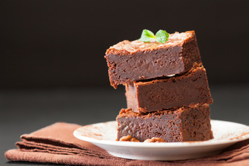 Chocolate brownie square pieces in stack on white plate with walnuts, decorated with mint leaves and cocoa on black background. Delicious dessert. Dark mood. Close up photography. Selective focus