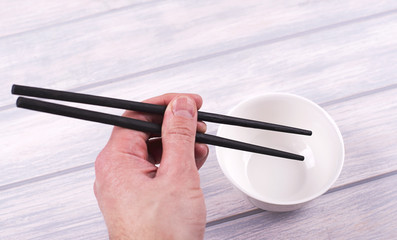 Hand holding Chinese chopsticks next to white bowl on wooden table.