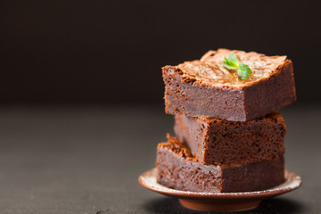 Chocolate brownie square pieces in stack on white plate with walnuts, decorated with mint leaves and cocoa on black background. Delicious dessert. Dark mood. Close up photography. Selective focus