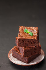 Chocolate brownie square pieces in stack on white plate with walnuts, decorated with mint leaves and cocoa on black background. Delicious dessert. Dark mood. Close up photography. Selective focus